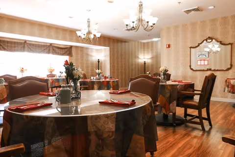 Dining room with round tables covered in patterned tablecloths, place settings and floral centerpieces under chandeliers on wood floors.