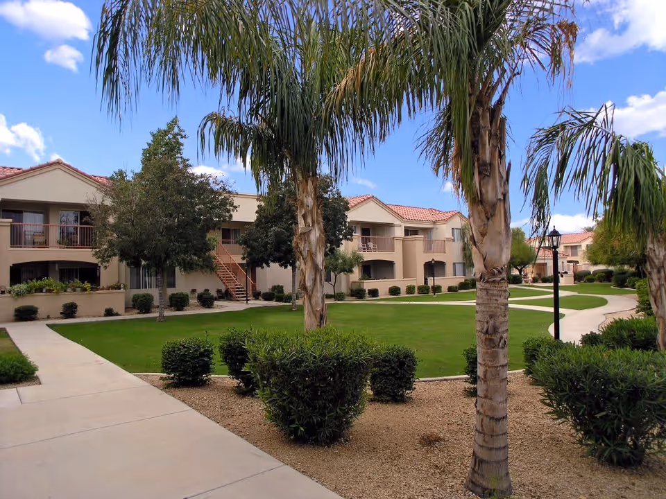 Outdoor view of a senior living facility courtyard with well-maintained green lawns, palm trees, shrubs, and paved walkways. Beige two-story buildings with red-tiled roofs and balconies surround the courtyard under a partly cloudy blue sky.