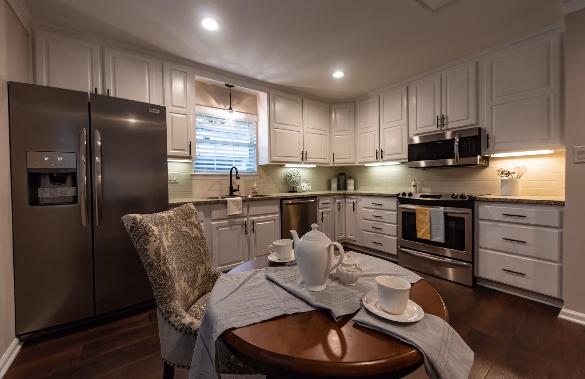 A modern kitchen with white cabinets, stainless steel appliances including a refrigerator, dishwasher, oven, and microwave. A small round wooden table is set with a white teapot, cups, and saucers, with a patterned upholstered chair beside it. The kitchen has dark wood flooring and under-cabinet lighting, with a window above the sink.