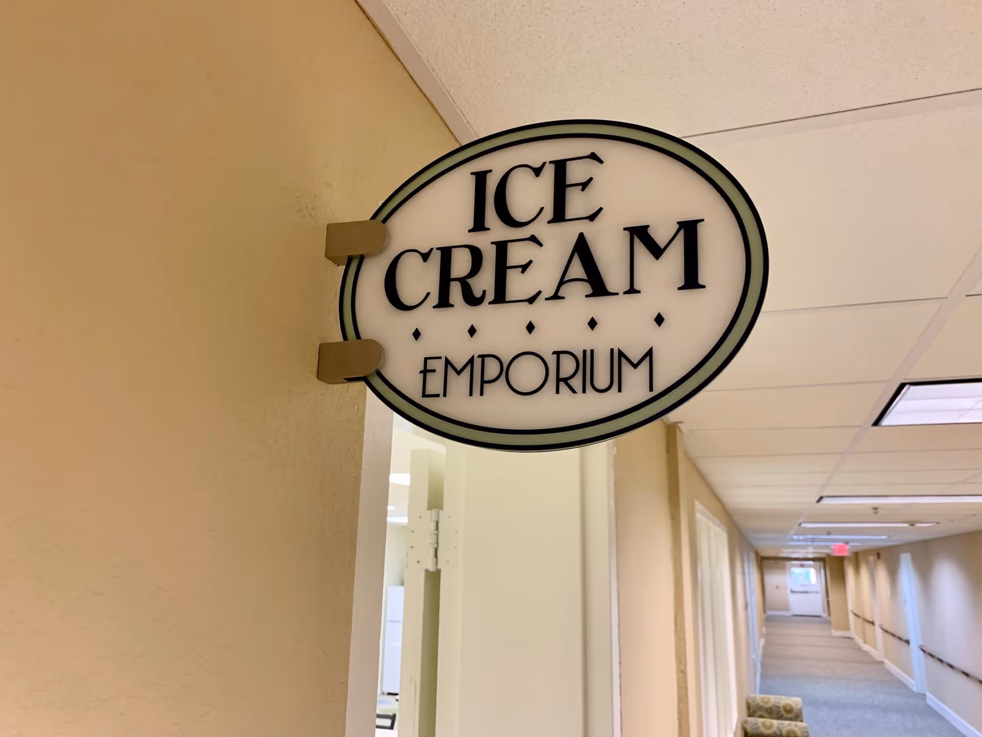 A hallway in a senior living facility with beige walls and ceiling tiles. A sign hanging from the wall reads 'ICE CREAM EMPORIUM'. The hallway has handrails and a chair is visible along the wall.