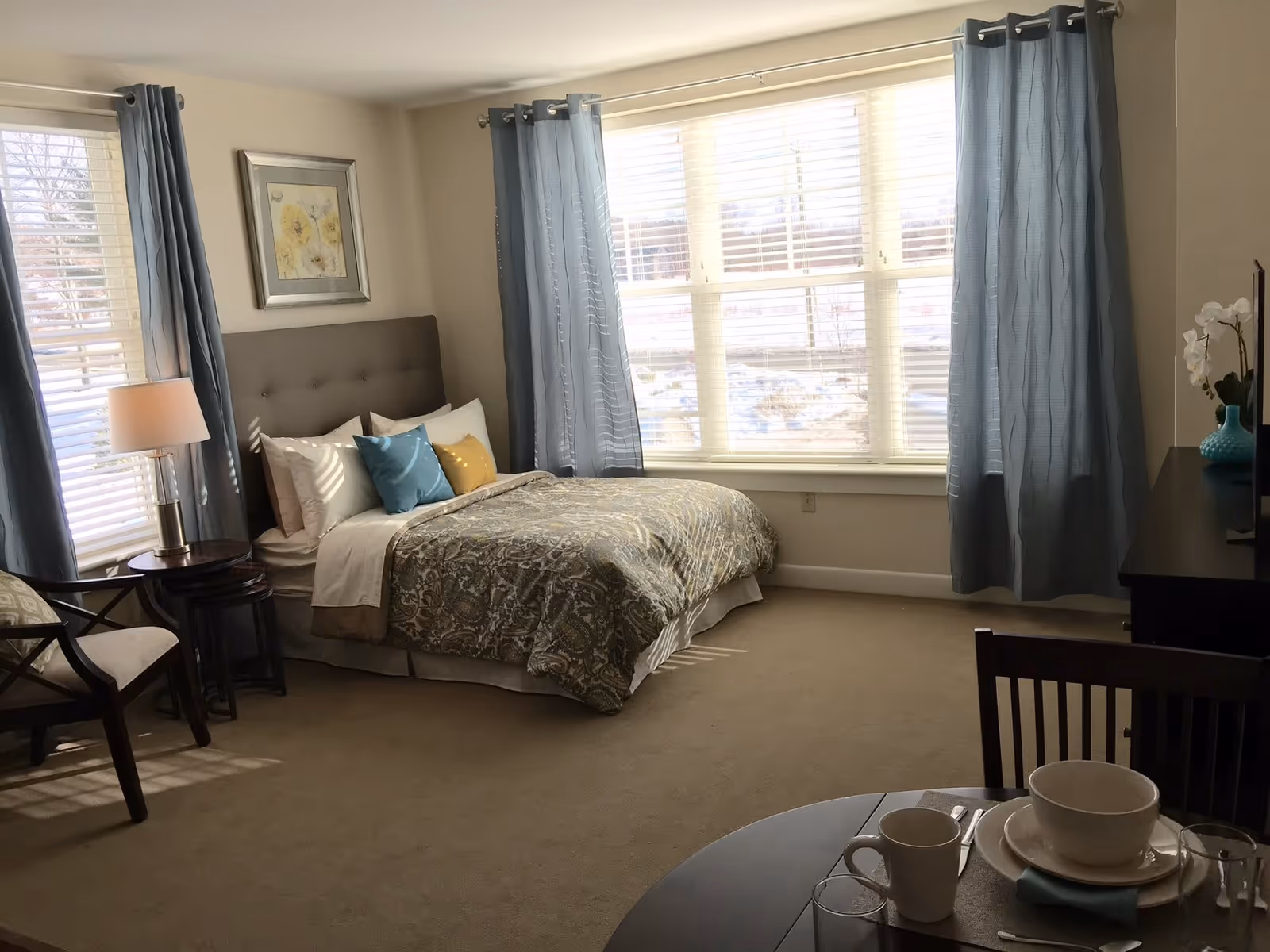 A bright bedroom with a large window covered by blue curtains. The room features a bed with a patterned comforter and several pillows, a nightstand with a lamp, a chair, and a small dining table set with dishes and cups. There is a framed floral picture on the wall above the bed and a dresser with a vase of flowers.