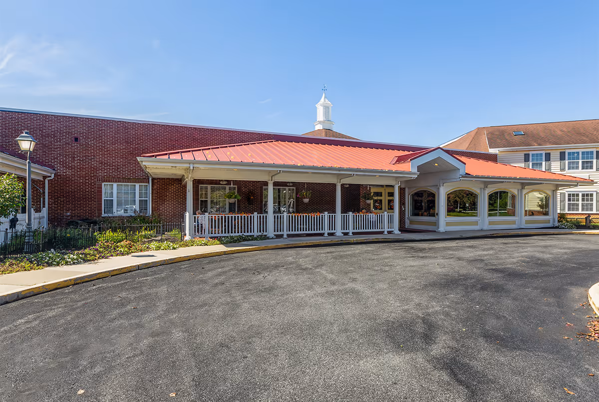 Front exterior view of Milford Place facility showing a brick building with a red metal roof, white railings, large windows, and a circular driveway under a clear blue sky.