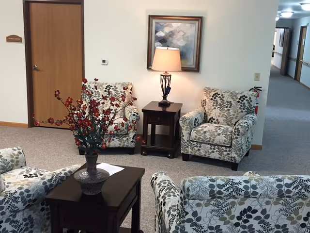 A cozy seating area in an assisted living facility with four armchairs upholstered in floral patterned fabric arranged around two dark wooden side tables. One table holds a vase with red flowers, and the other has a lamp with a beige shade. A framed abstract painting hangs on the wall behind the lamp. A hallway with doors and ceiling lights extends to the right.
