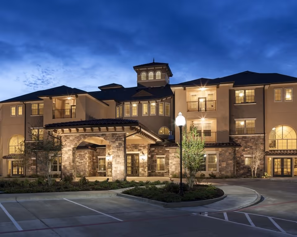 Exterior view of a multi-story senior living facility named Trinity Courtyard at dusk, featuring warm lighting, stone and stucco facade, a covered entrance, balconies, and a well-maintained driveway with parking spaces and landscaping.