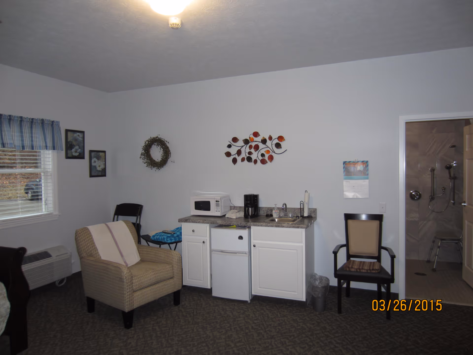 Interior room with a small kitchenette featuring a microwave, coffee maker, sink, and mini fridge. There is a beige armchair with a towel draped over it, a chair with a cushion, and wall decorations including a wreath and a metal leaf art piece. A window with plaid curtains is on the left, and an open door on the right reveals a bathroom with grab bars and a shower chair.