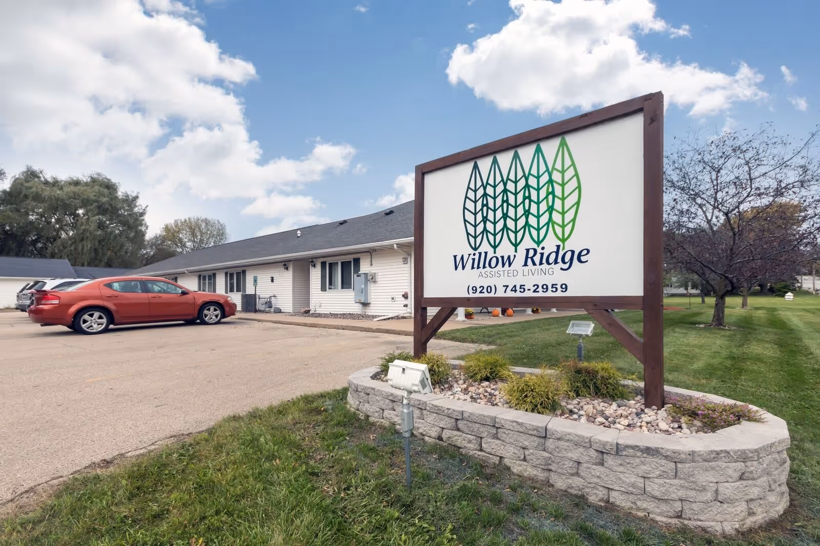 Exterior view of Willow Ridge Assisted Living facility with a large sign displaying the facility name and phone number. The building is a single-story structure with white siding and a gray roof. There are a few parked cars in the parking lot, green grass, trees, and a partly cloudy sky.