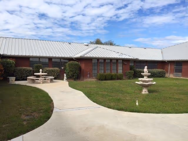Outdoor courtyard area of a senior living facility with a concrete pathway, green grass, a stone fountain, and a stone picnic table with benches. The building has a white metal roof and red brick walls with windows and bushes along the walls. The sky is partly cloudy.