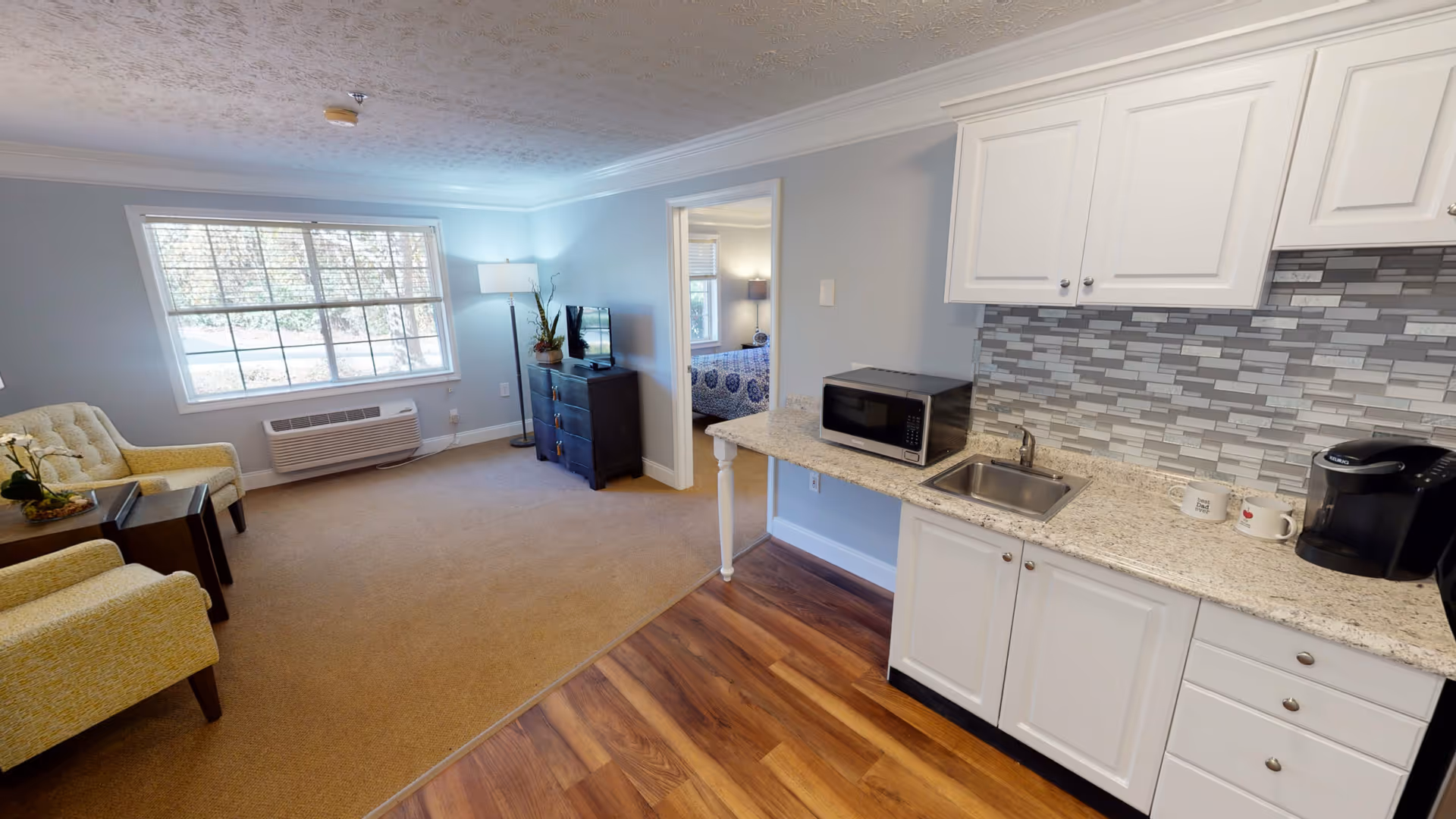 Bright studio-style living room with seating by a large window and an adjacent kitchenette with white cabinets and a microwave.
