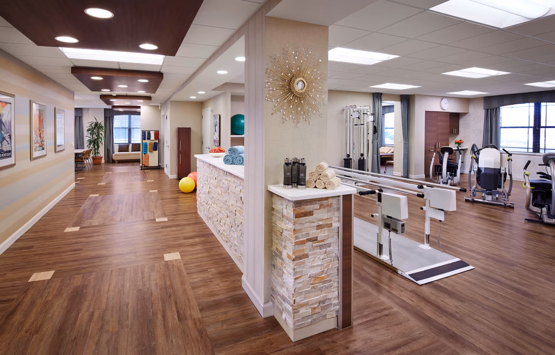 Interior view of a senior living facility showing a hallway with wooden flooring and a fitness area on the right equipped with exercise machines, parallel bars, and rolled towels on a counter. The hallway has framed artwork on the walls, recessed ceiling lights, and a potted plant near a window at the end.