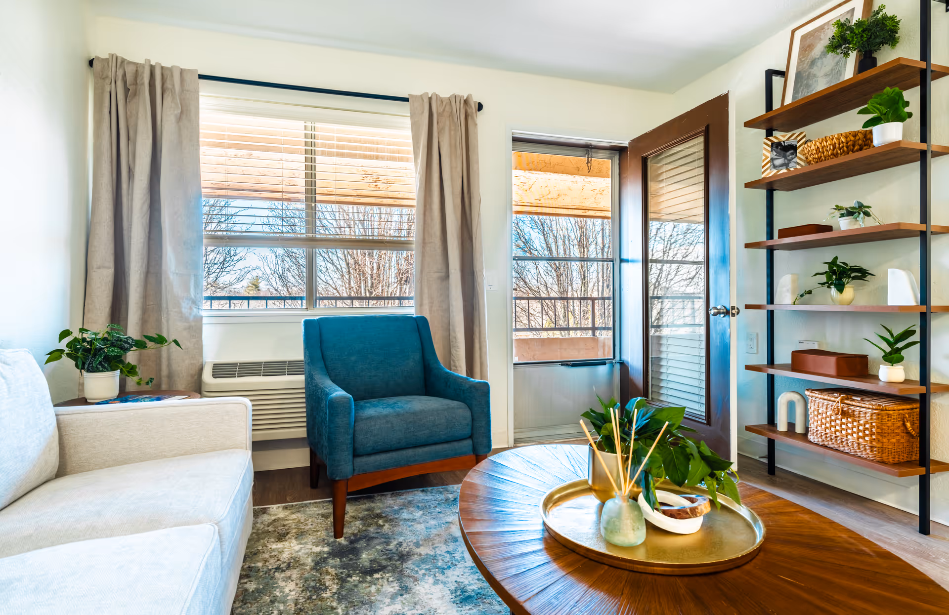 A bright and cozy living room with a beige sofa on the left, a blue armchair near a window with beige curtains, and a wooden round coffee table with decorative plants and a tray. There is a door leading to an outdoor balcony and a wooden shelving unit with various plants and decorative items on the right.