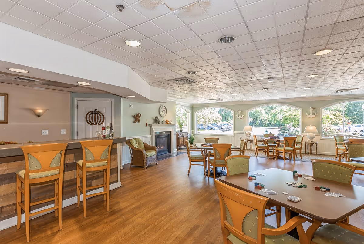 A bright and spacious common area with wooden floors, multiple tables and chairs arranged for socializing or playing games, a bar counter with two high chairs, a cozy fireplace, large windows letting in natural light, and decorative wall anchors.