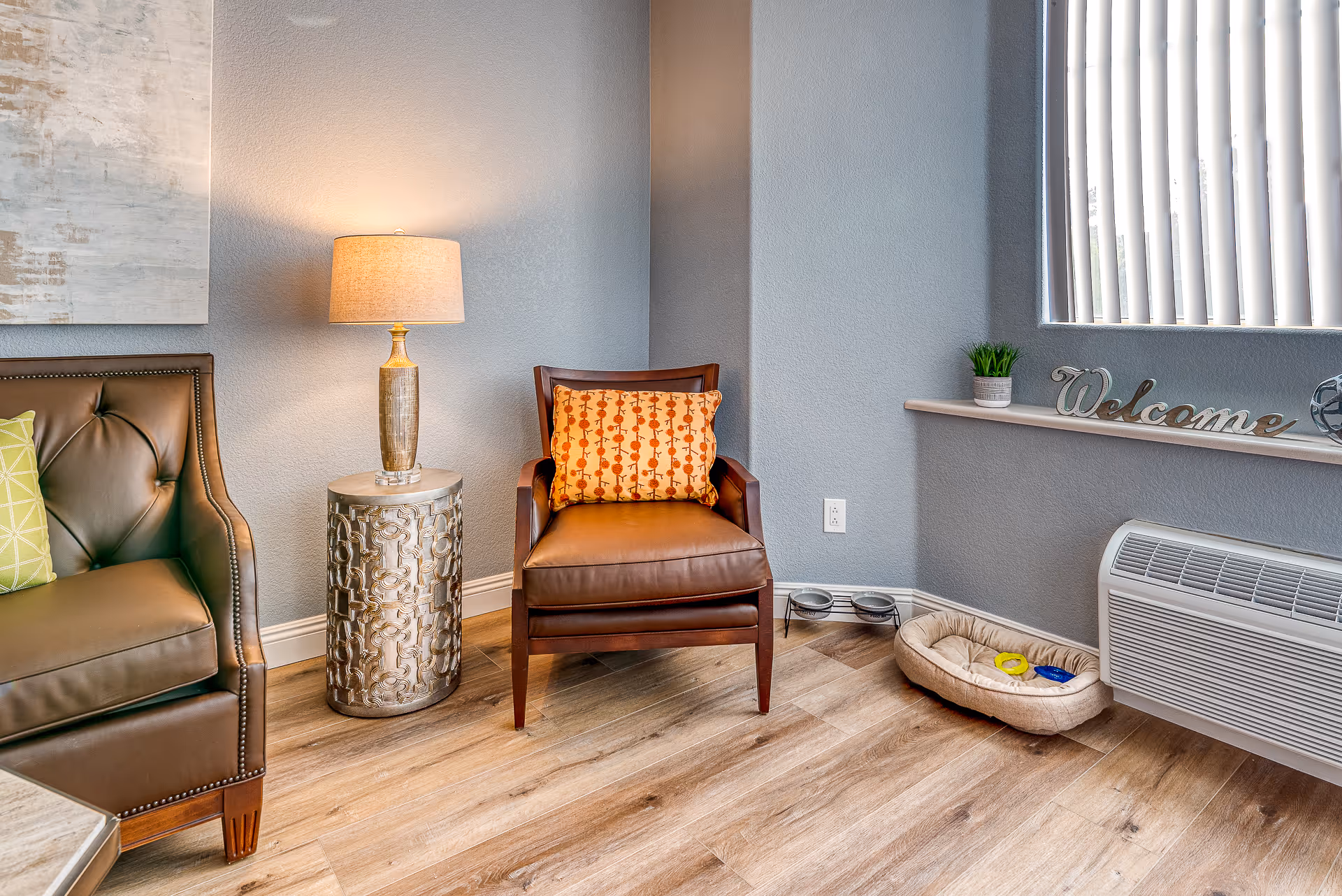 A cozy corner of a living room with a brown leather armchair featuring an orange patterned cushion, a decorative silver side table with a lamp, a brown leather sofa with a green pillow, a pet bed with toys, a small plant, and a decorative 'Welcome' sign on the windowsill with vertical blinds.