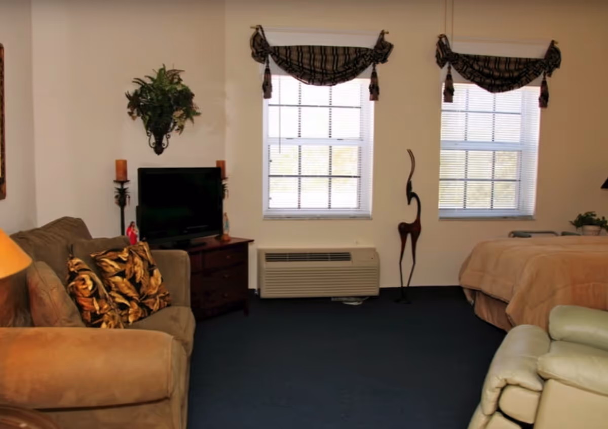 Studio-style living area with a sofa and armchair, TV on a dresser, and a bed next to two windows with valances.