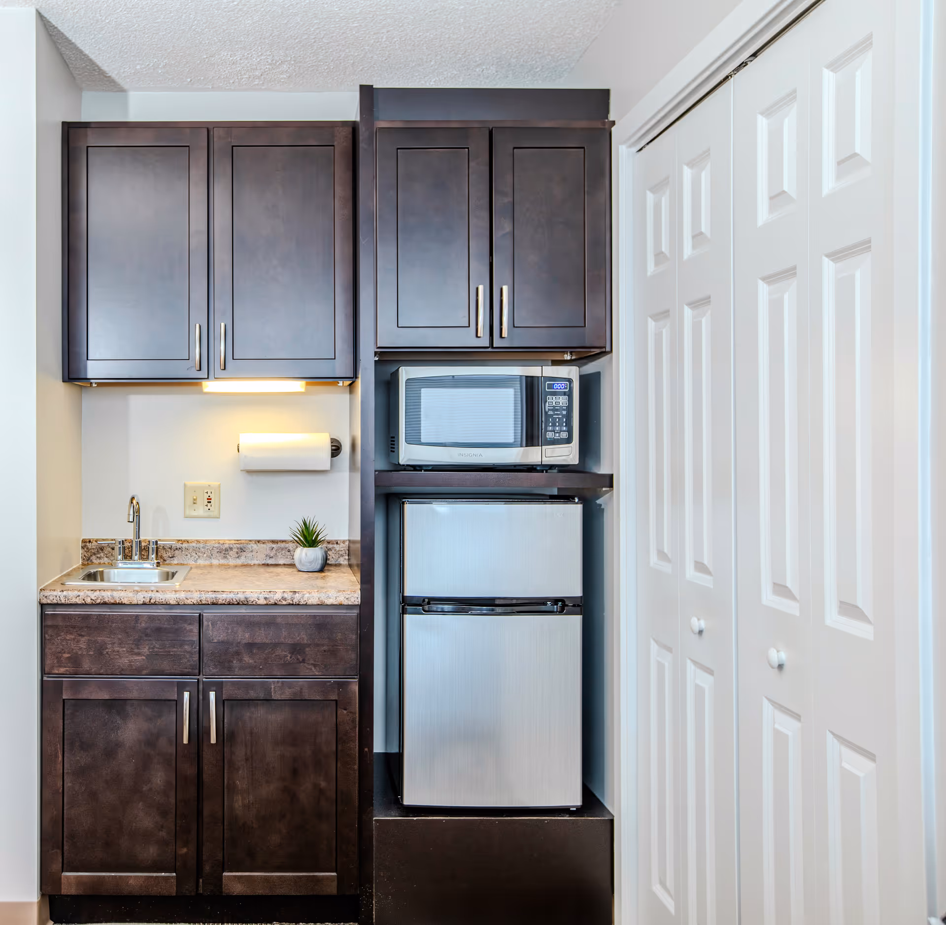 Small kitchenette with dark wood cabinets, a sink, a microwave above a mini refrigerator, and white bi-fold closet doors.