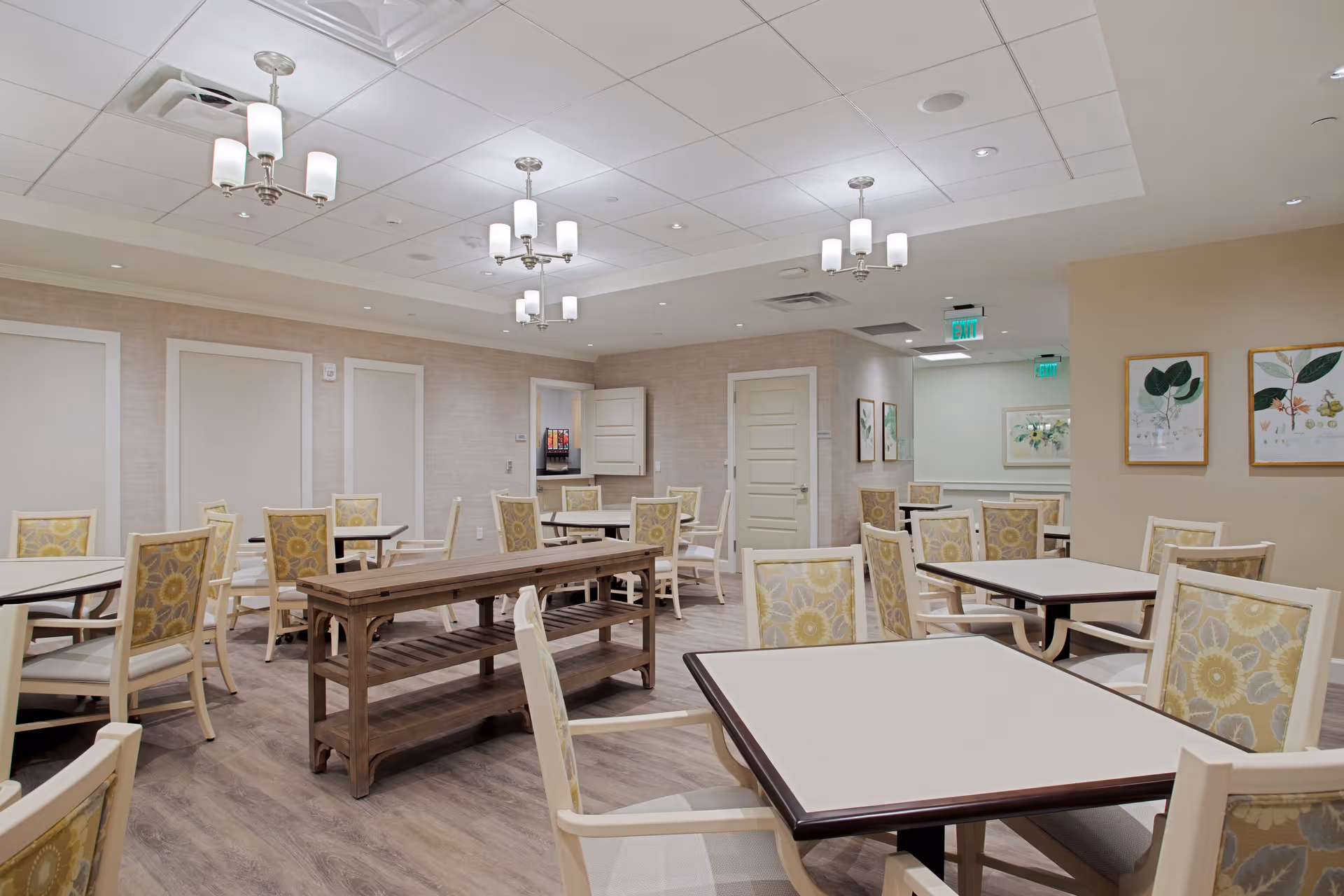A spacious dining room with multiple tables and chairs featuring floral-patterned upholstery. The room has a light-colored wood floor, beige walls, and several ceiling light fixtures. There are framed botanical prints on one wall and a wooden bench-style table in the center. Doors and an exit sign are visible in the background.