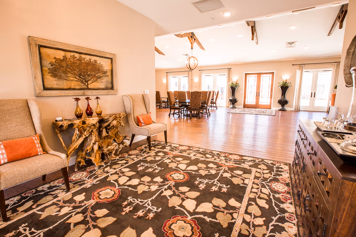 Spacious, well-lit senior community lounge with armchairs, a decorative console and rug, a dining table in the background, and double glass doors.