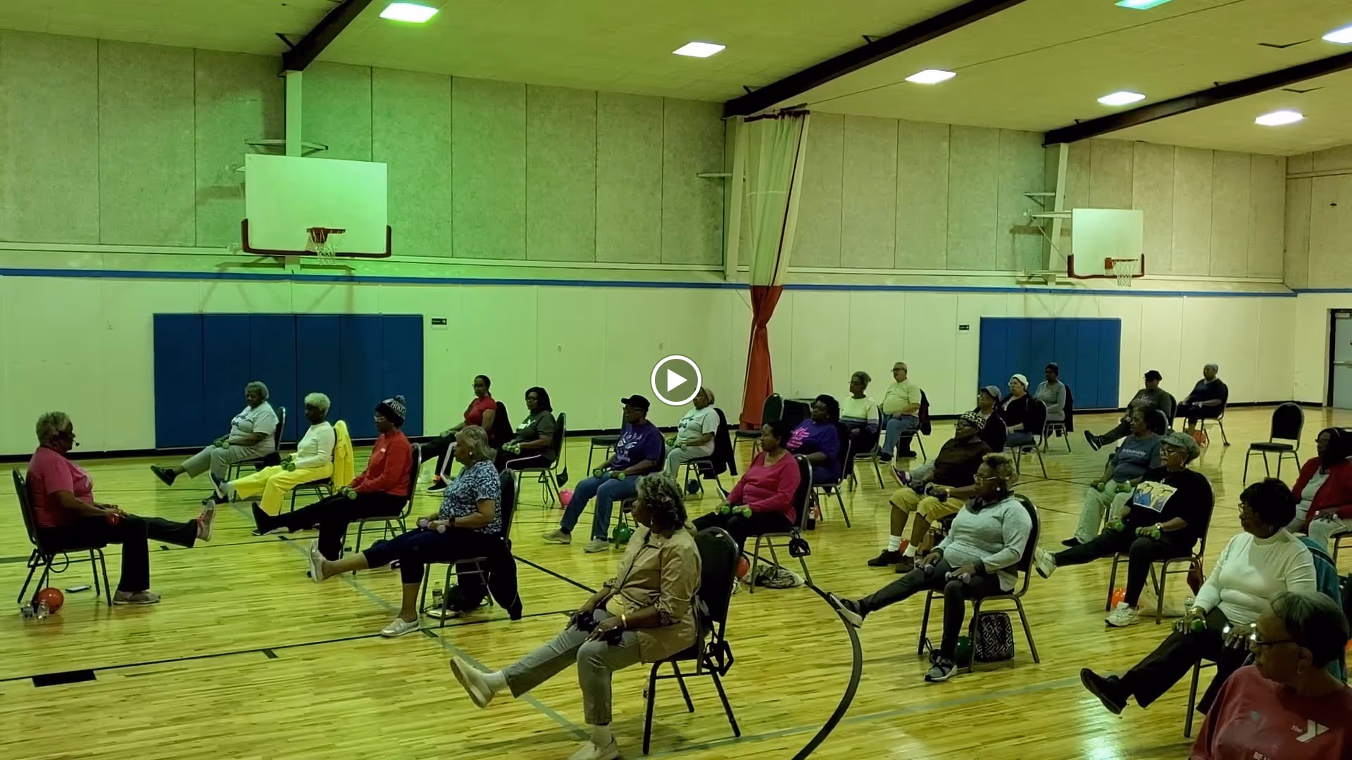 A group of elderly individuals seated on chairs in a gymnasium participating in a seated exercise class, extending one leg forward. The gym has wooden floors, basketball hoops, and padded walls.