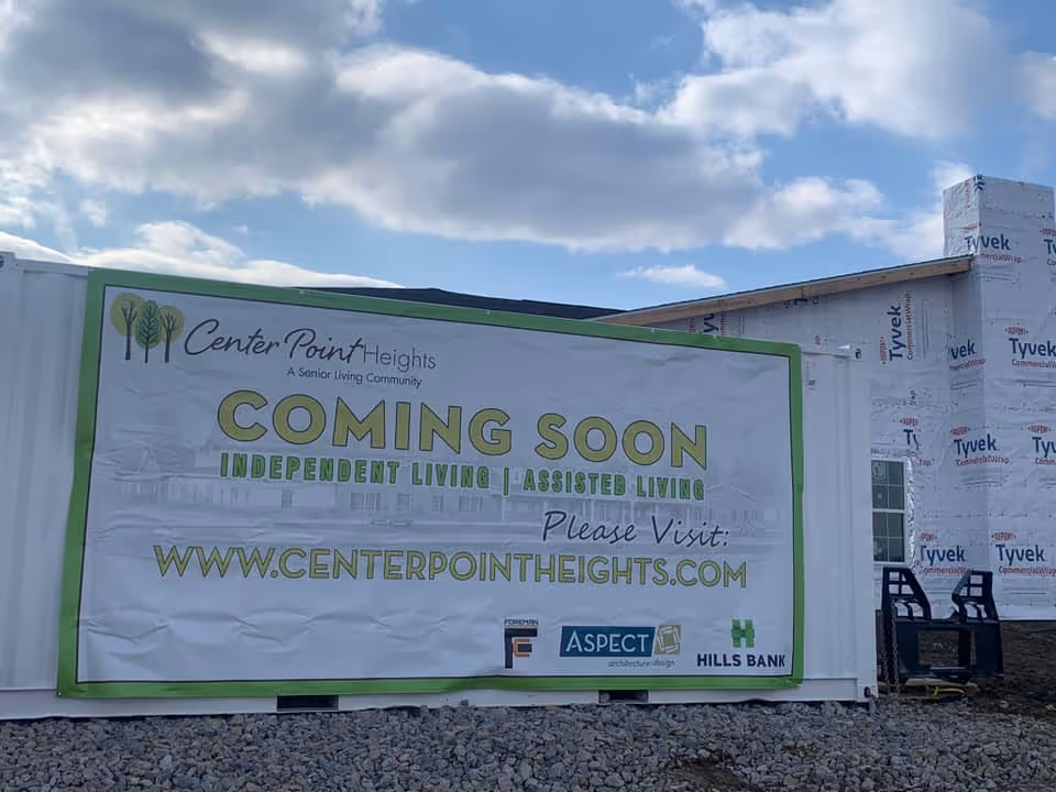 A construction site with a large white banner attached to a container. The banner reads 'Center Point Heights A Senior Living Community COMING SOON INDEPENDENT LIVING | ASSISTED LIVING Please Visit: WWW.CENTERPOINTHEIGHTS.COM'. The building under construction is wrapped in Tyvek material. The sky is partly cloudy.