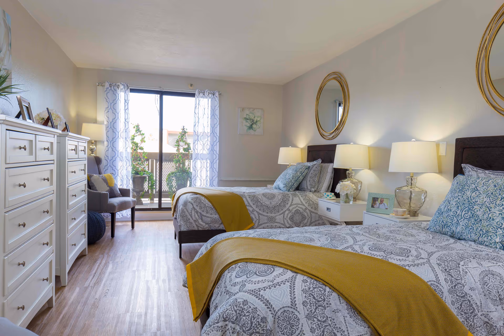 A bright and neatly arranged bedroom with two twin beds, each adorned with patterned bedding and yellow throws. The room features wooden flooring, two white dressers on the left, a comfortable chair with cushions, and a sliding glass door leading to a balcony with plants. Two round mirrors and bedside lamps are positioned above the nightstands between the beds.