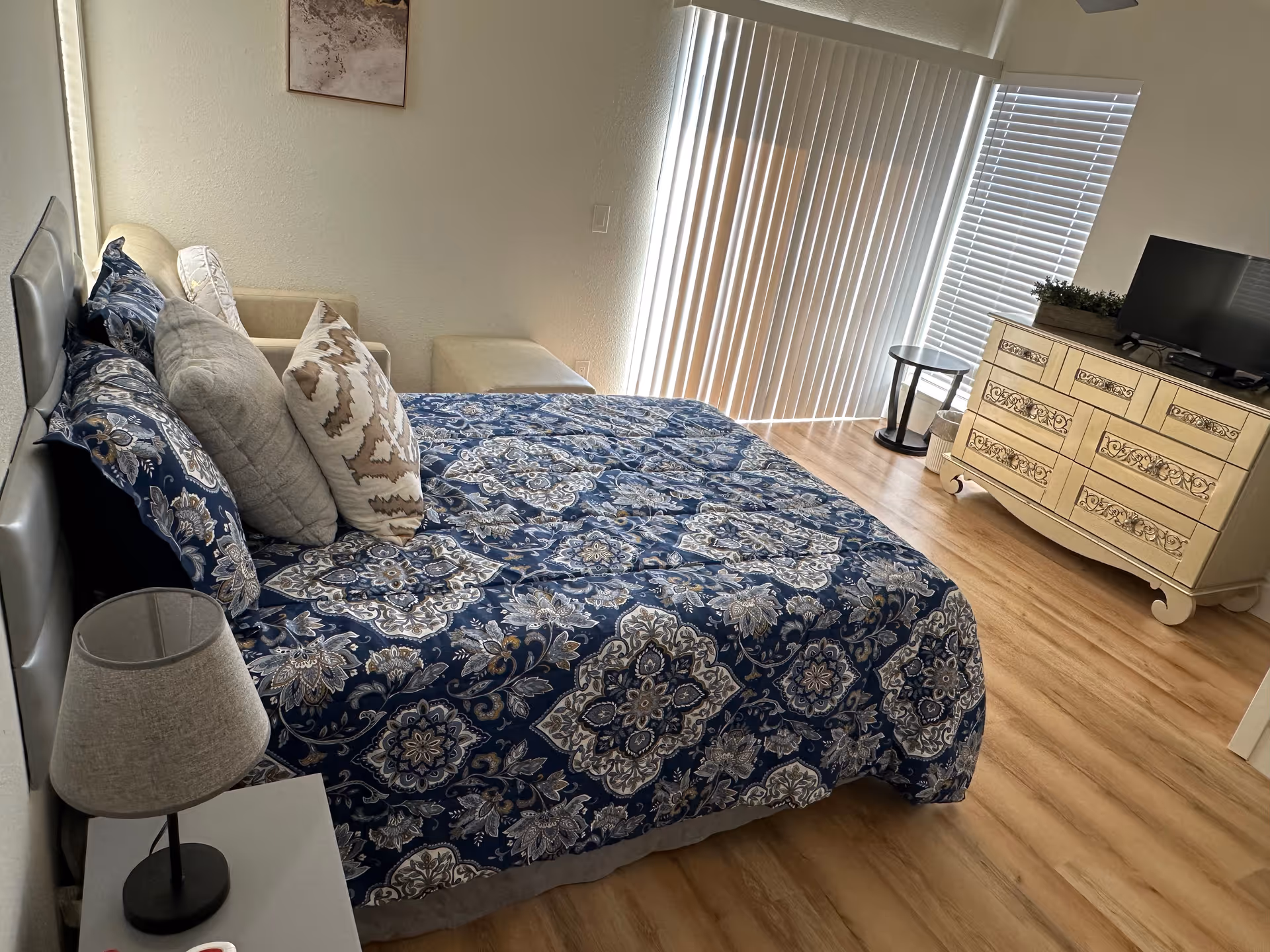 A bedroom with a bed covered in a blue and white patterned bedspread, several pillows, a beige armchair with an ottoman, a small round side table, a cream-colored dresser with decorative drawer fronts, a flat-screen TV on top of the dresser, vertical blinds covering a sliding glass door, and a window with horizontal blinds.