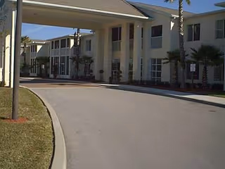 Exterior view of a two-story senior living facility building with a covered driveway entrance, palm trees, and a clear blue sky.