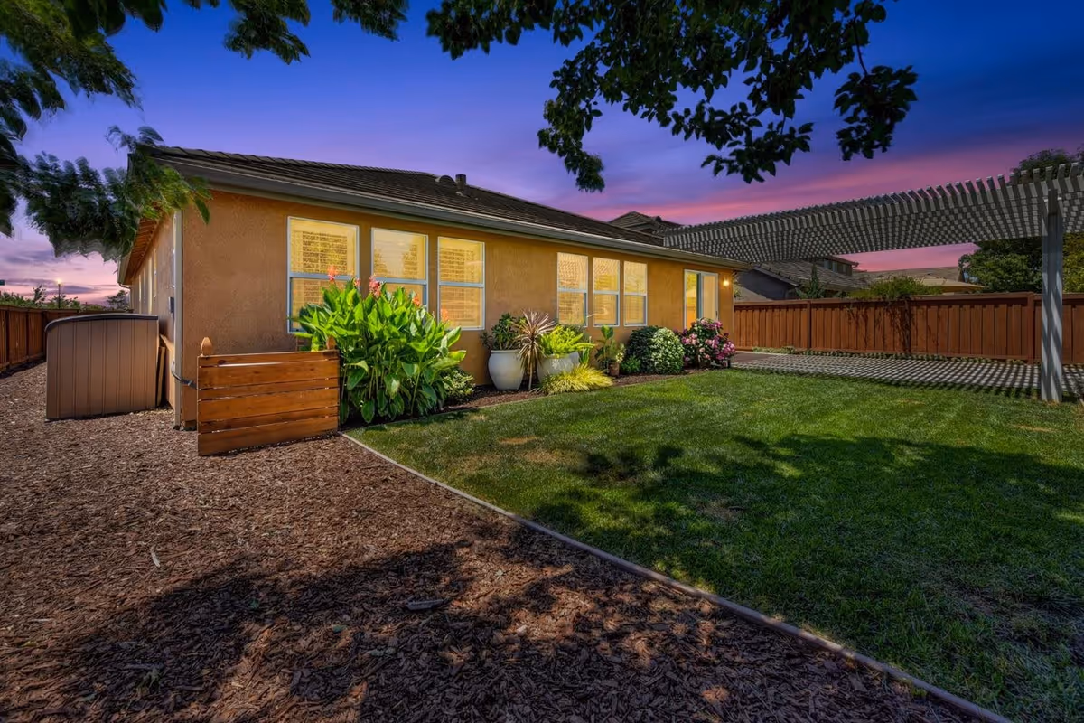 A backyard view of a single-story house at dusk with a well-maintained lawn, various plants and flowers along the house wall, a wooden fence surrounding the yard, and a pergola casting shadows on the grass.