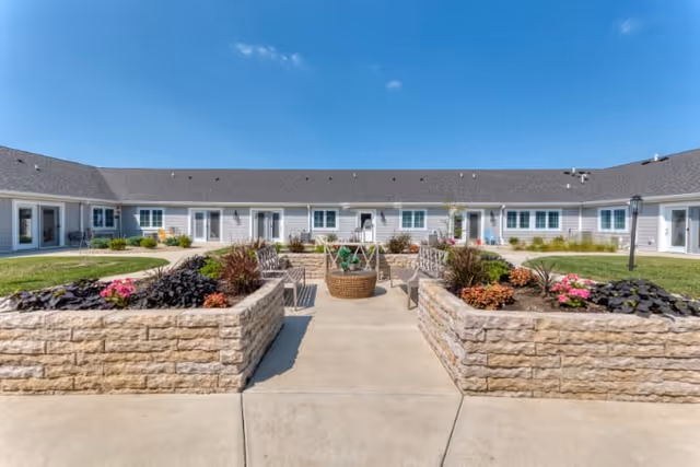 Outdoor courtyard area at Cedarhurst Senior Living of Springfield featuring raised stone flower beds with colorful plants and flowers, surrounded by a paved walkway and seating benches, with a single-story building in the background under a clear blue sky.