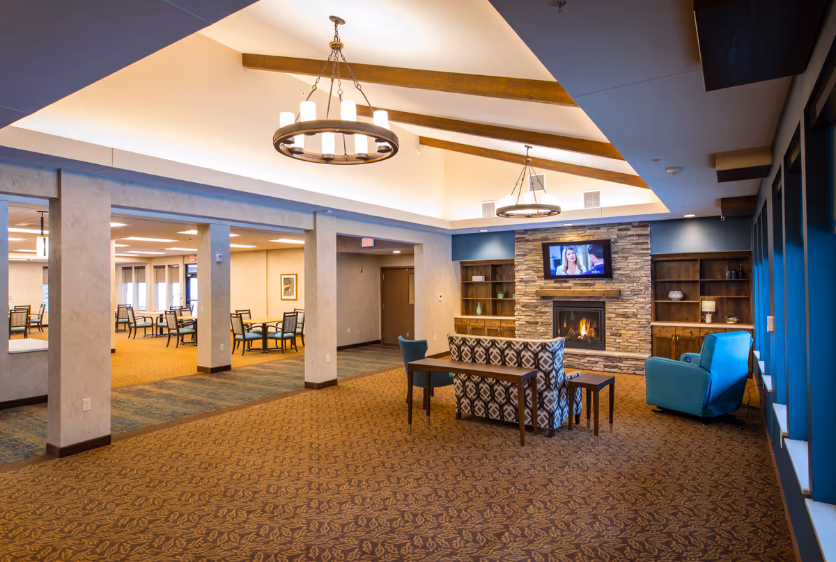 Spacious senior living common area with seating, a stone fireplace and wall-mounted TV, chandeliers, and an adjacent dining area.