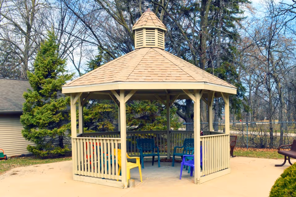 A wooden octagonal gazebo with colorful plastic chairs inside set on a paved patio surrounded by trees.