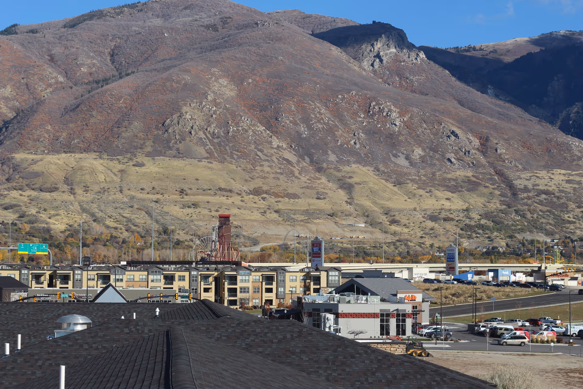View of a commercial area with buildings, parking lots, and a road in front of a large mountain with sparse vegetation under a clear blue sky.