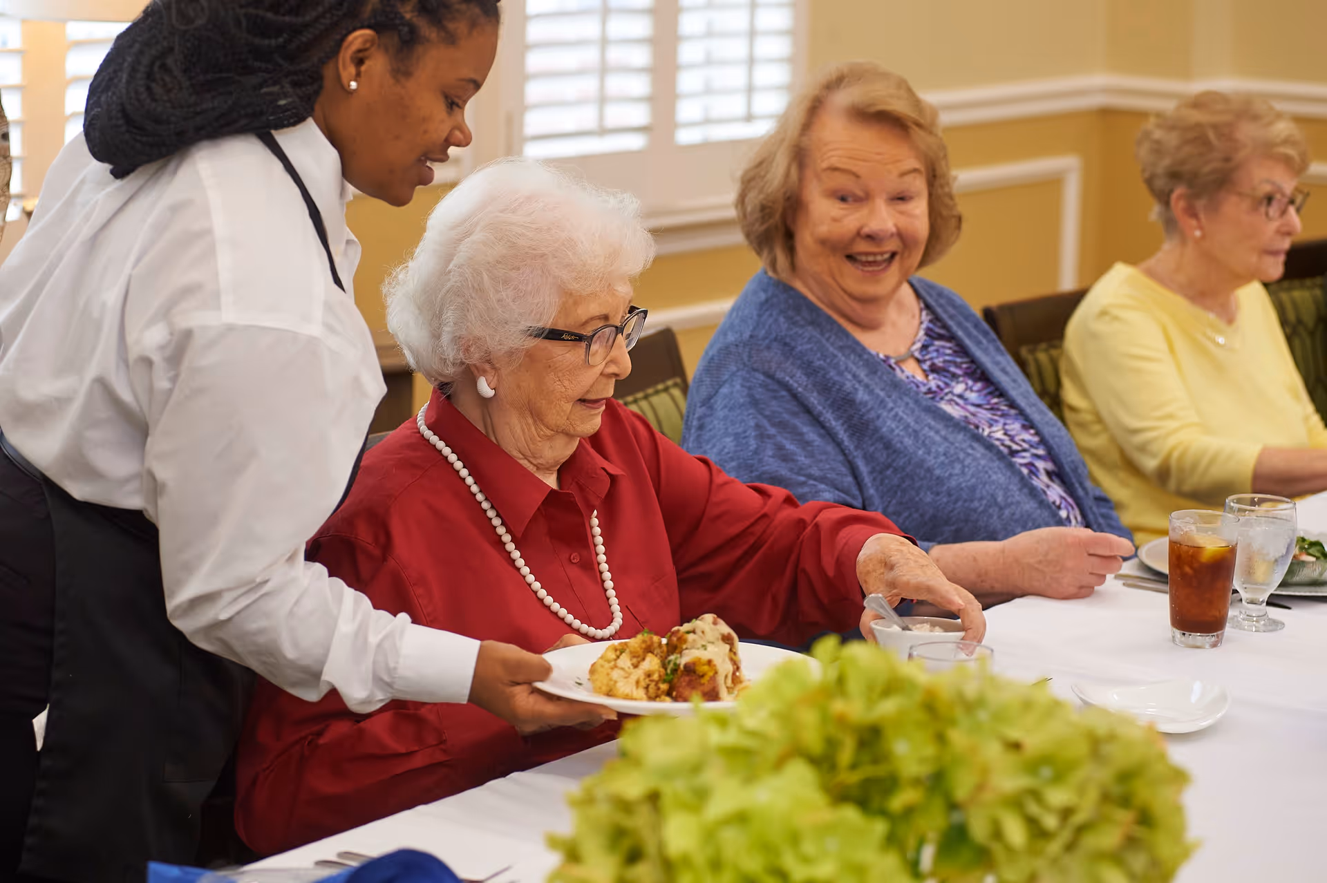 A caregiver serving a plate of food to an elderly woman wearing glasses and a red blouse seated at a dining table with two other elderly women, one in a blue cardigan and the other in a yellow top. The table is set with drinks and plates, and there is a green floral centerpiece in the foreground.
