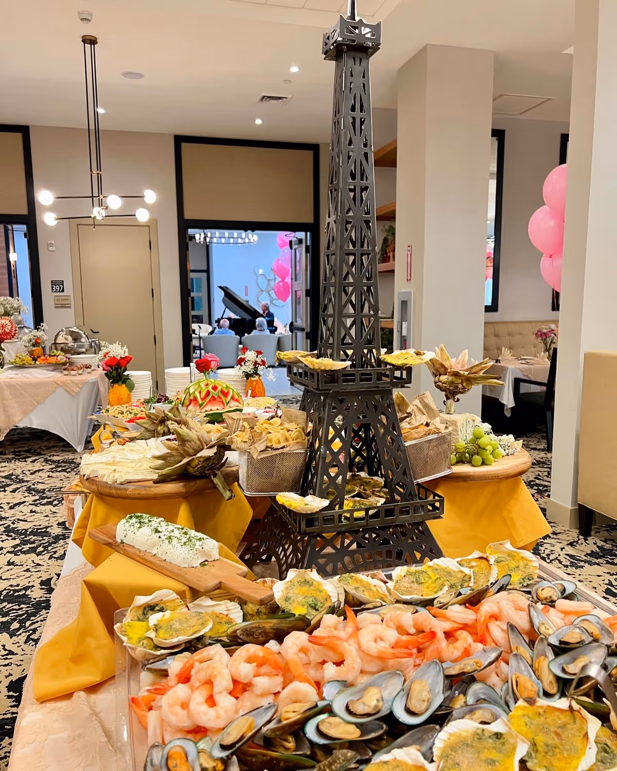 A buffet table set up with a variety of seafood including shrimp, mussels, and baked oysters, along with other appetizers and garnishes. The centerpiece is a decorative black metal Eiffel Tower. The room has a patterned carpet, modern lighting fixtures, and tables with white tablecloths and pink balloons in the background.