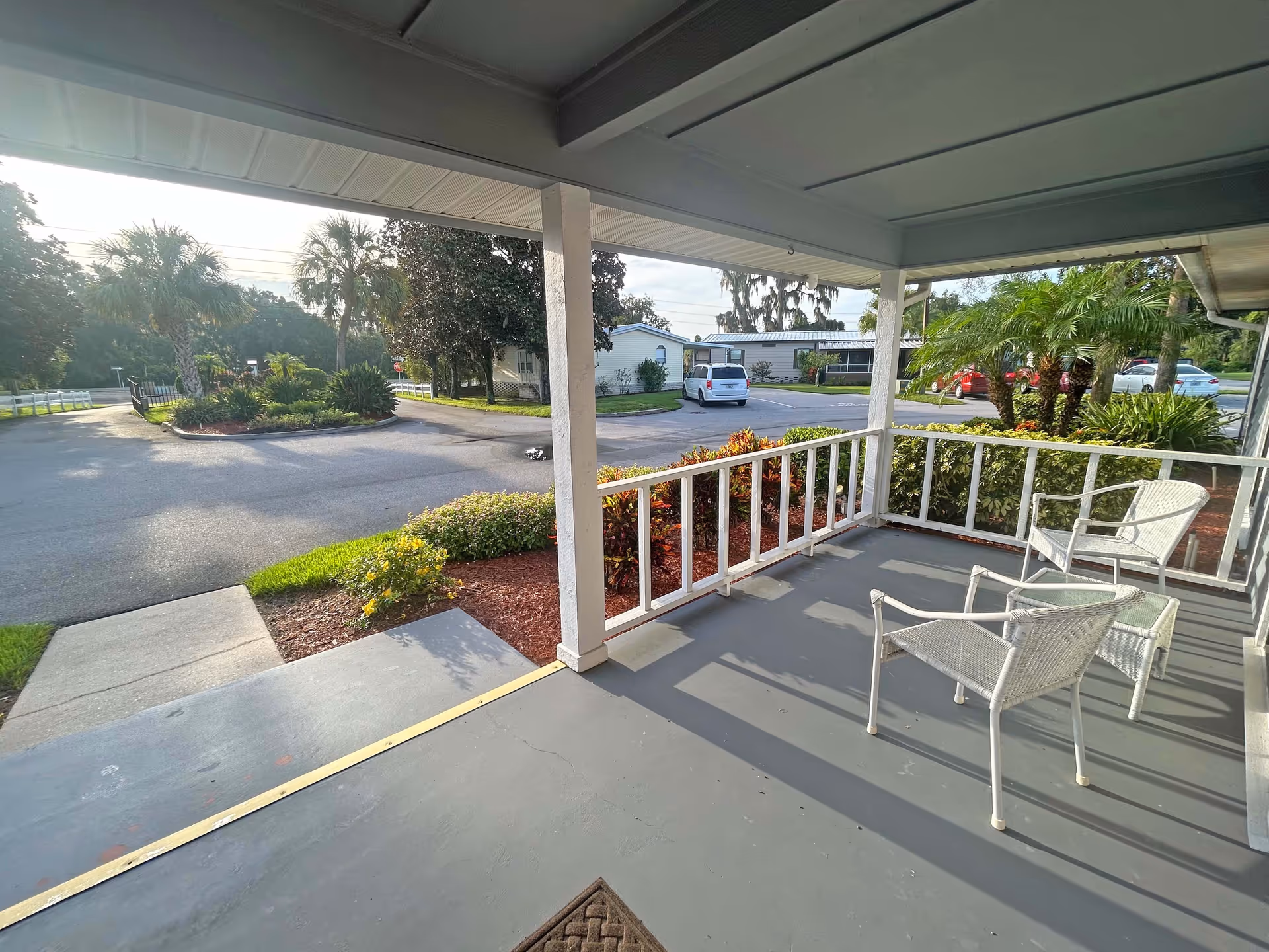 Covered porch area with two white wicker chairs and a small table, overlooking a parking lot with several cars and landscaped greenery including palm trees and bushes.