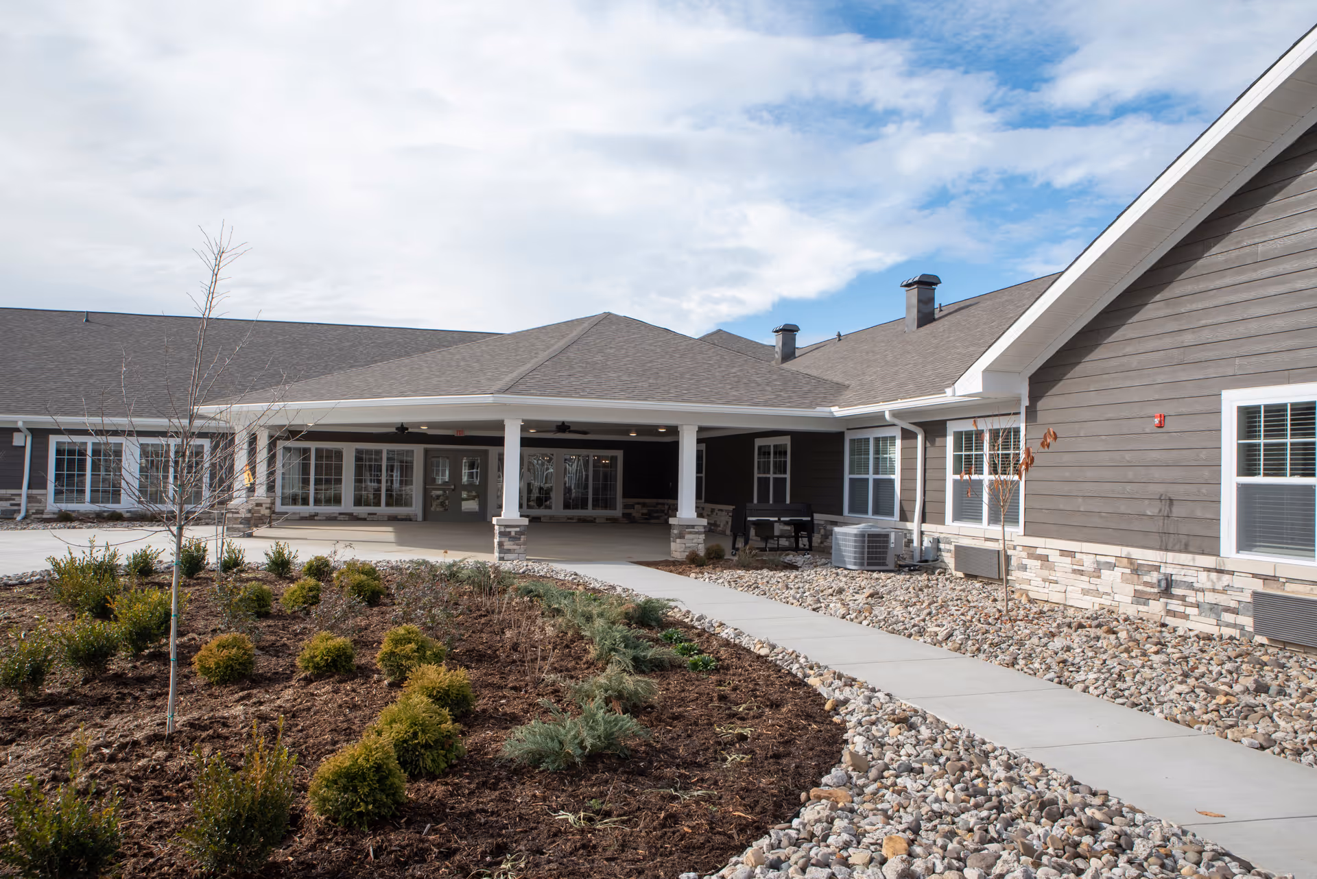 Front exterior of a single-story senior living building with a covered entrance, paved walkway, and landscaped beds.