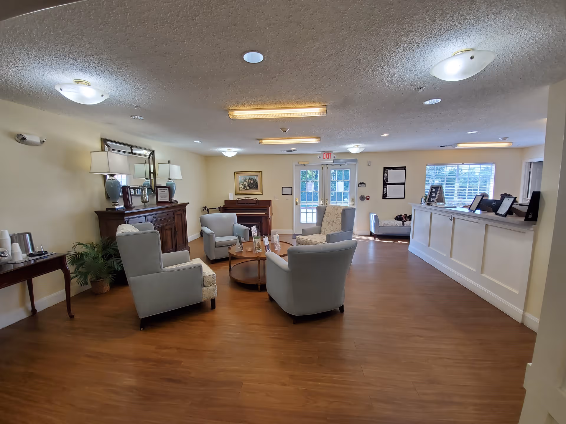 A spacious senior living facility common area with wooden flooring, four upholstered armchairs arranged around a circular wooden coffee table, a wooden sideboard with lamps and decorative items, a piano against the far wall, and a reception desk on the right side near a window and double glass doors.