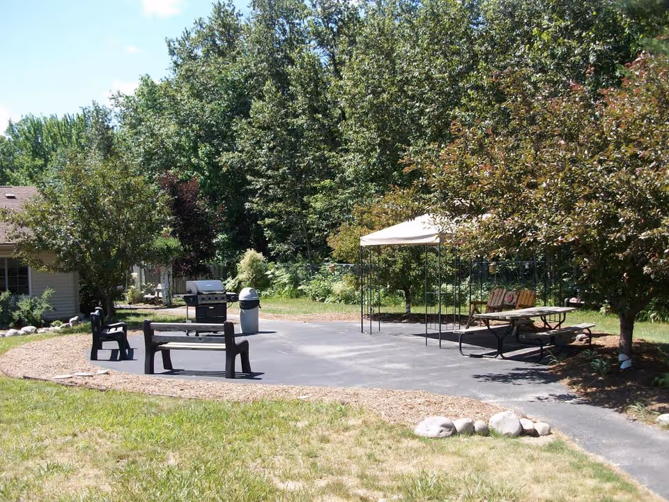 Paved outdoor patio with benches, a grill, a canopy-covered picnic table, and surrounding trees.