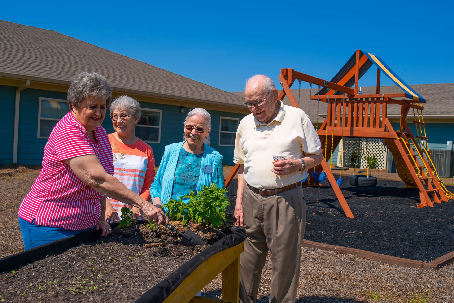 Four elderly people gardening together outdoors near a raised garden bed with plants. They are smiling and enjoying the sunny day. In the background, there is a playground structure with swings and a climbing wall, and a blue building with windows.