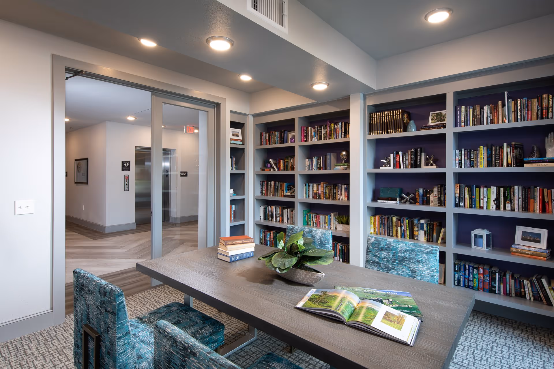 Bright communal library/reading room with wall bookshelves, a table with chairs, and an open book on the table.