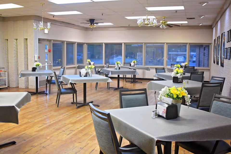 Spacious dining room with multiple square tables covered in gray tablecloths, chairs, and floral centerpieces beside large windows.