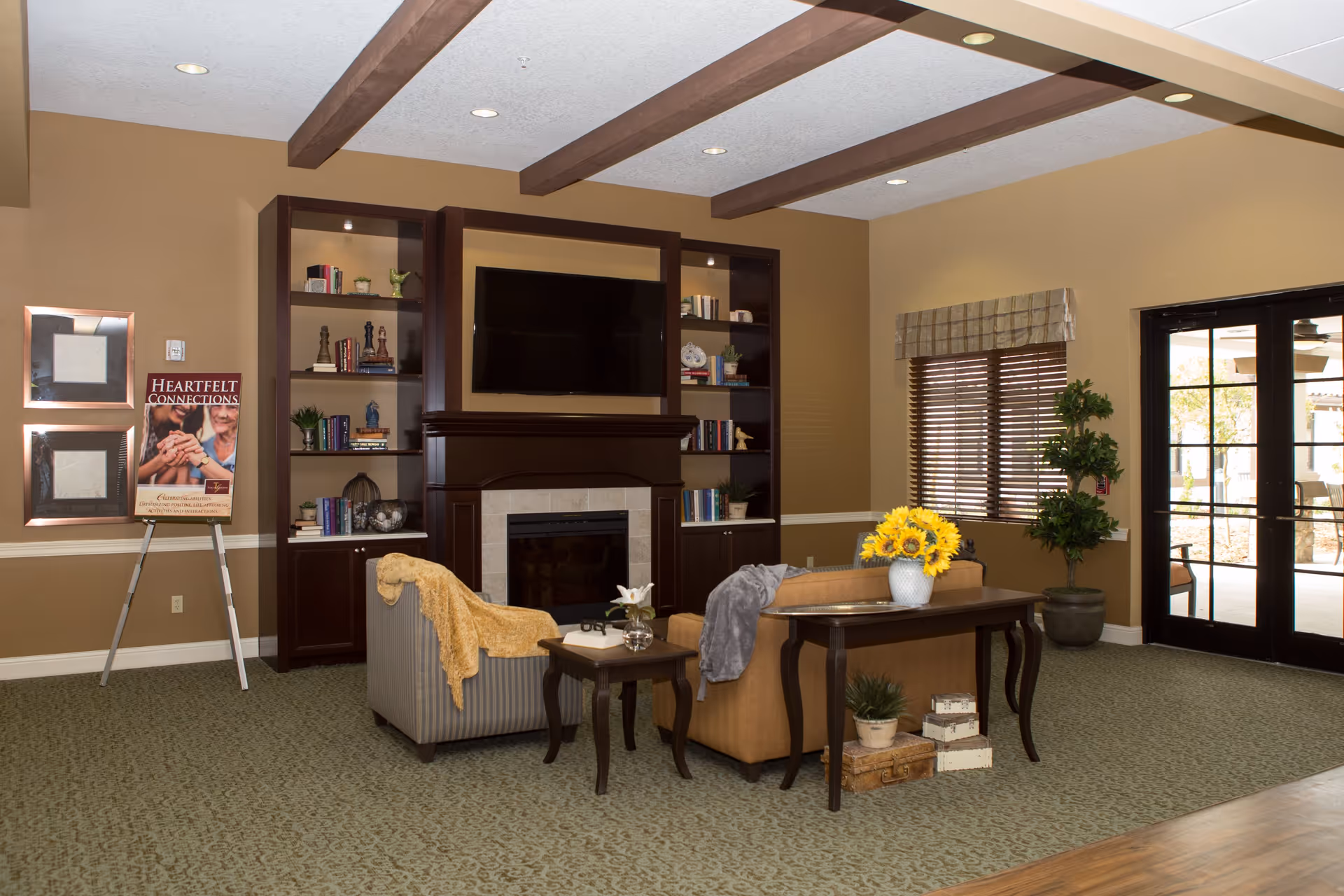 A cozy living room area in Tuscan Gardens Of Venetia Bay featuring a fireplace with built-in dark wood shelves filled with books and decorative items. Two armchairs with throws are arranged around a small table with a flower vase. A larger table behind one chair holds a vase with sunflowers and some decorative boxes underneath. The room has beige walls, a patterned carpet, wooden ceiling beams, and a window with blinds next to glass double doors leading outside.