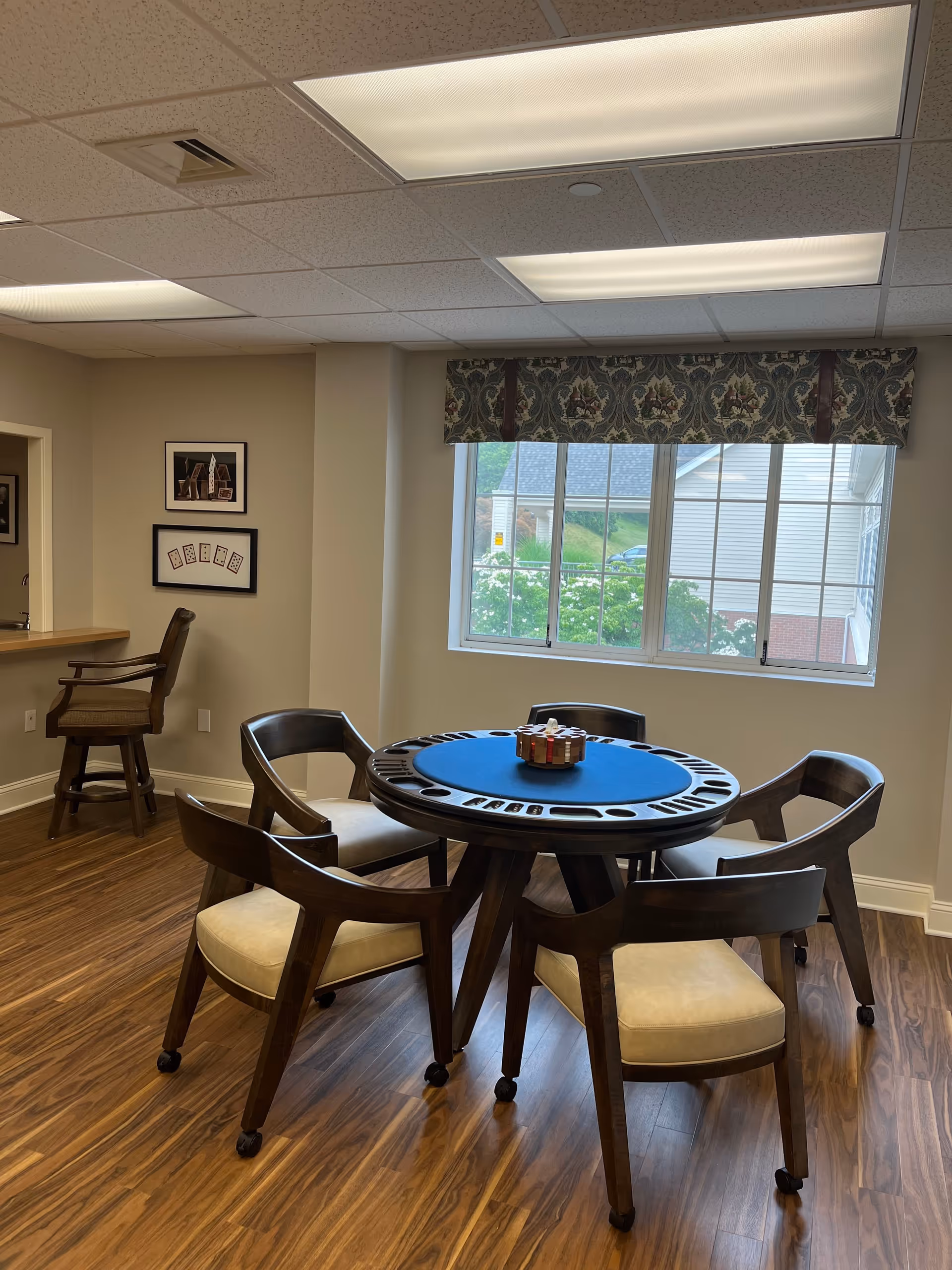 A well-lit room with a round card table covered with a blue felt top and surrounded by five wooden chairs with beige cushions on wheels. The room has wood flooring, a window with a floral valance, and two framed pictures on the wall. A high chair is positioned near a counter on the left side.