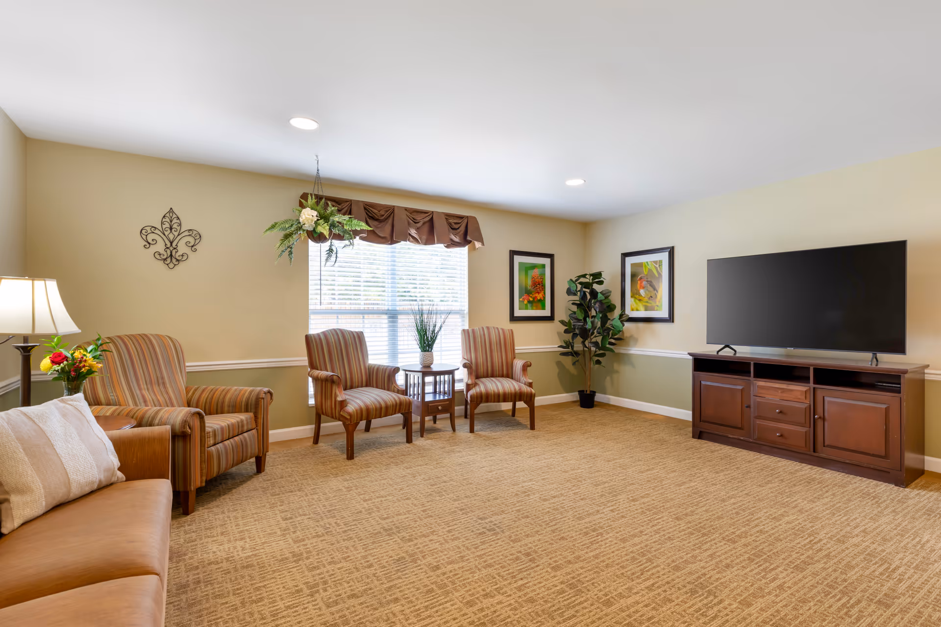 A cozy living room with beige walls and carpeted floor. The room features a brown leather sofa with a white pillow, two striped armchairs, a small wooden side table with a plant, a floor lamp with a bouquet of flowers, a large flat-screen TV on a wooden TV stand, two framed pictures on the wall, a hanging plant near the window with blinds, and a potted plant in the corner.