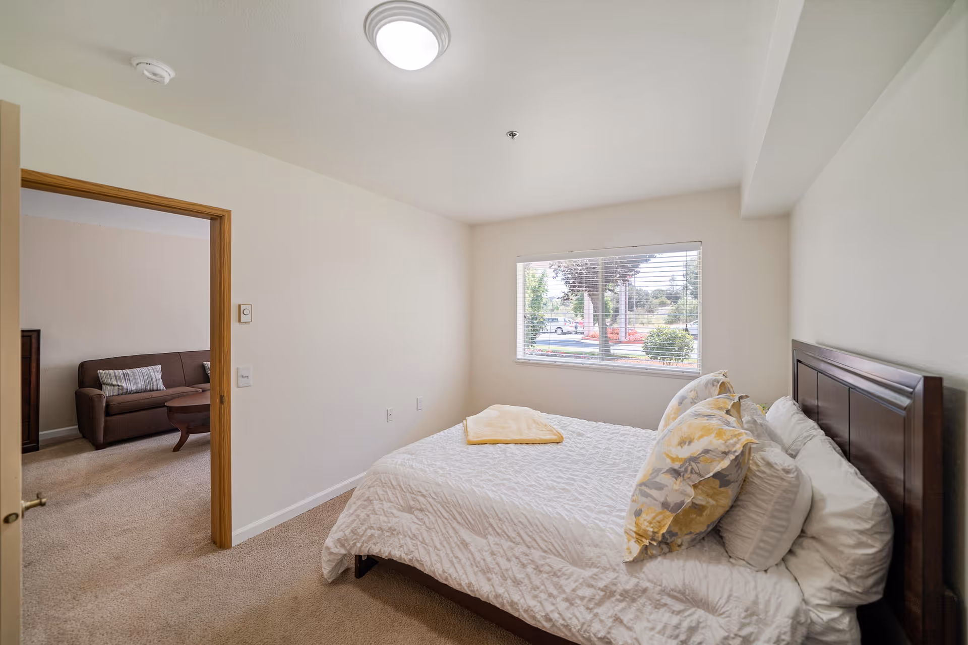 A bedroom with a neatly made bed featuring white bedding and floral pillows. There is a window with blinds letting in natural light, and an open door leading to a living area with a brown couch and a wooden coffee table.