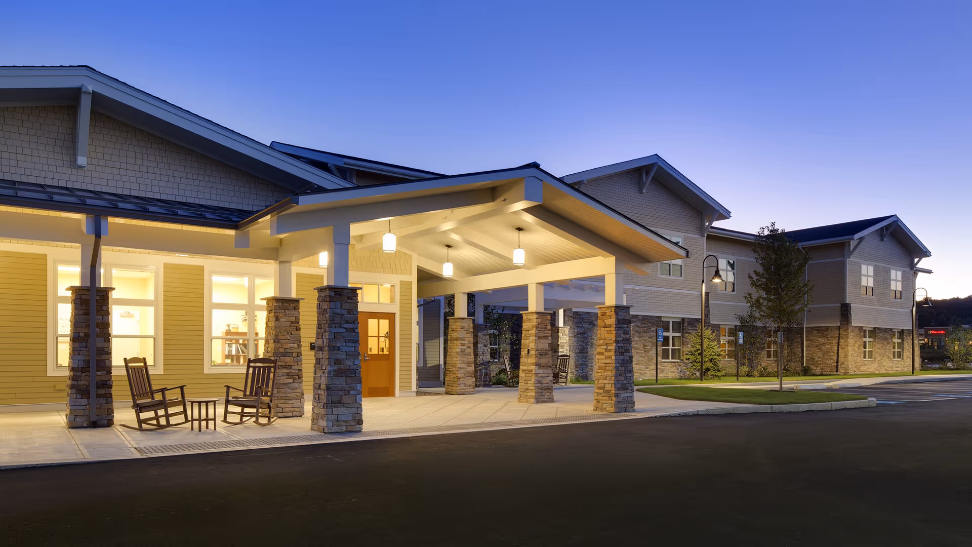 Front entrance of a senior living facility at dusk featuring a covered porte‑cochere with stone columns and rocking chairs.