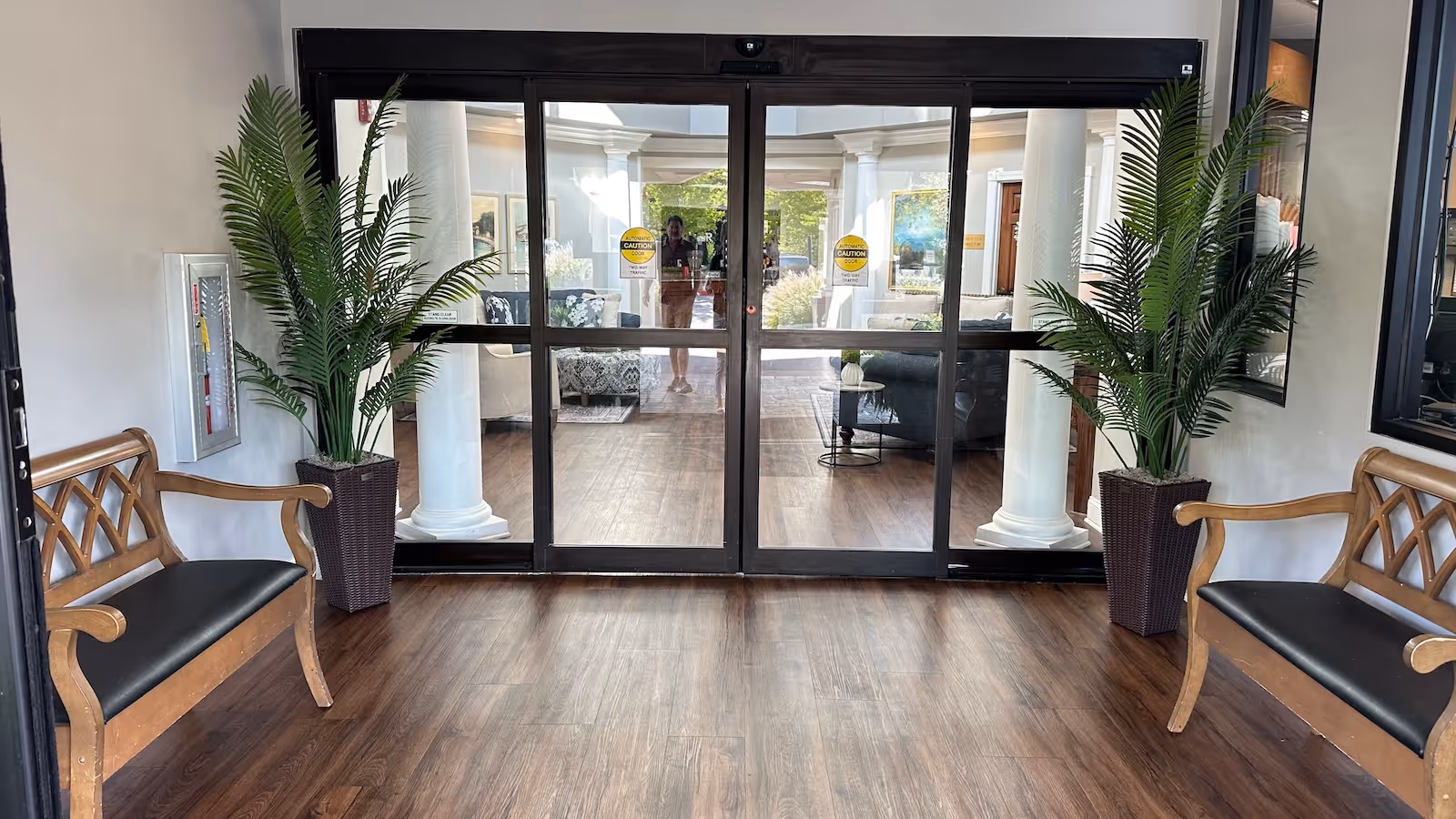 Entrance lobby with two wooden benches, potted plants, and sliding glass doors opening into a furnished seating area.