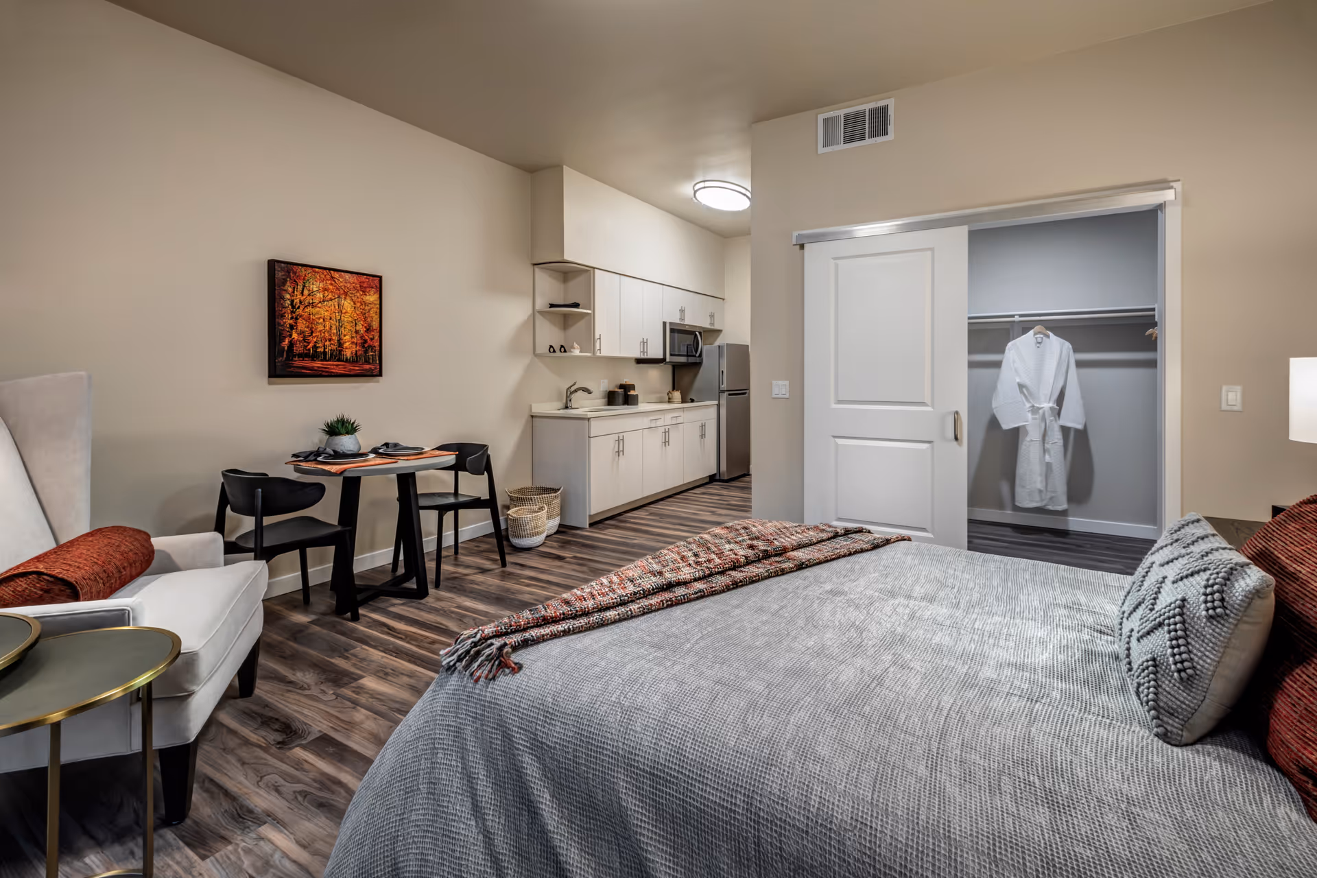 A cozy studio apartment featuring a bed with gray bedding and decorative pillows, a white armchair with a red throw blanket, a small dining table with two black chairs, and a kitchenette with white cabinets, a sink, microwave, and refrigerator. A closet with a sliding door is open, showing a white robe hanging inside. The room has wood flooring and a framed picture of autumn trees on the wall.
