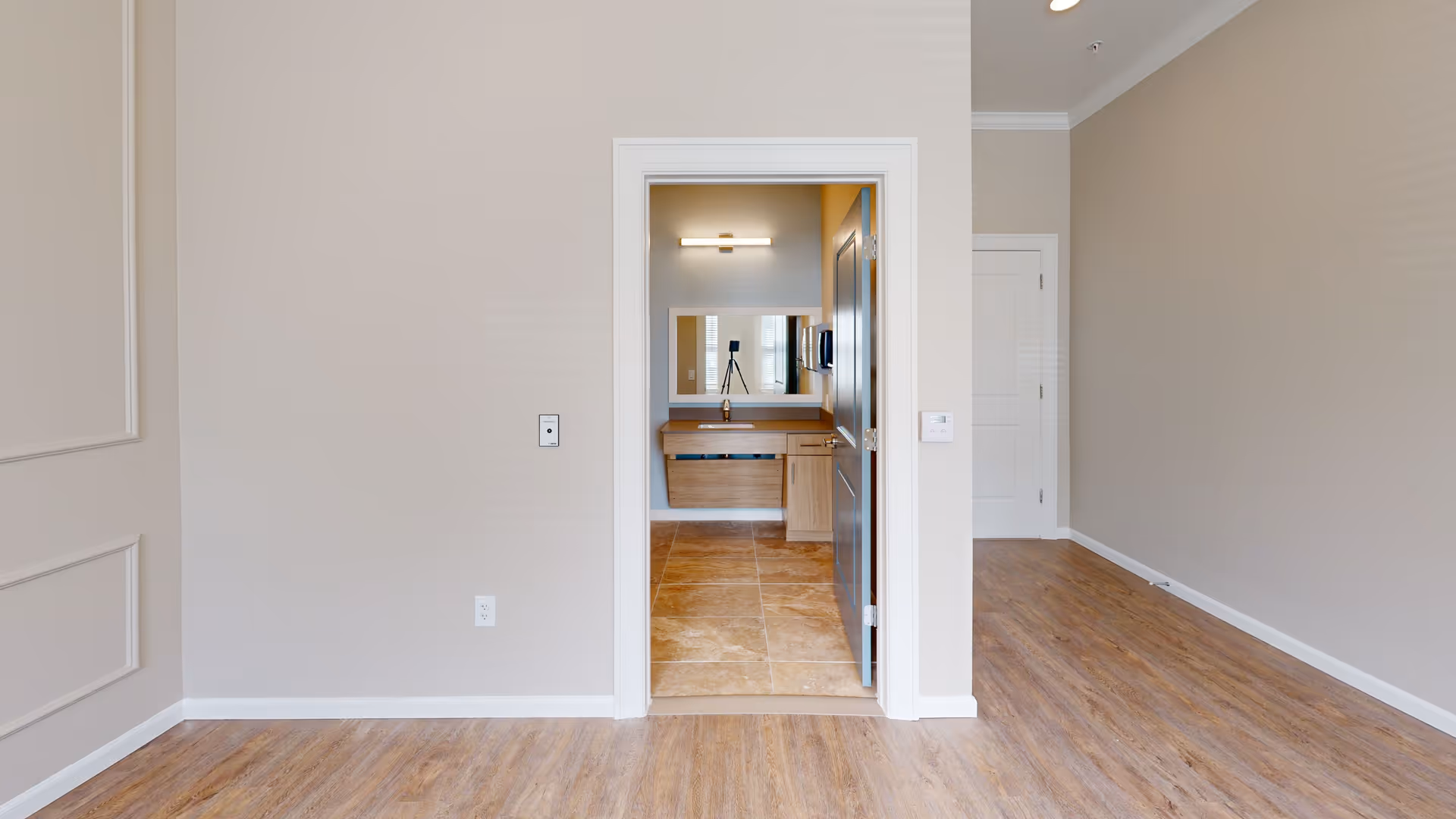 Empty interior room with wood-look flooring and an open doorway revealing a bathroom vanity and mirror.
