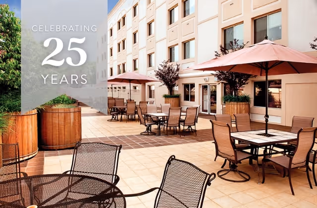 Sunny outdoor courtyard with patio tables, chairs, and umbrellas next to a multi-story building.
