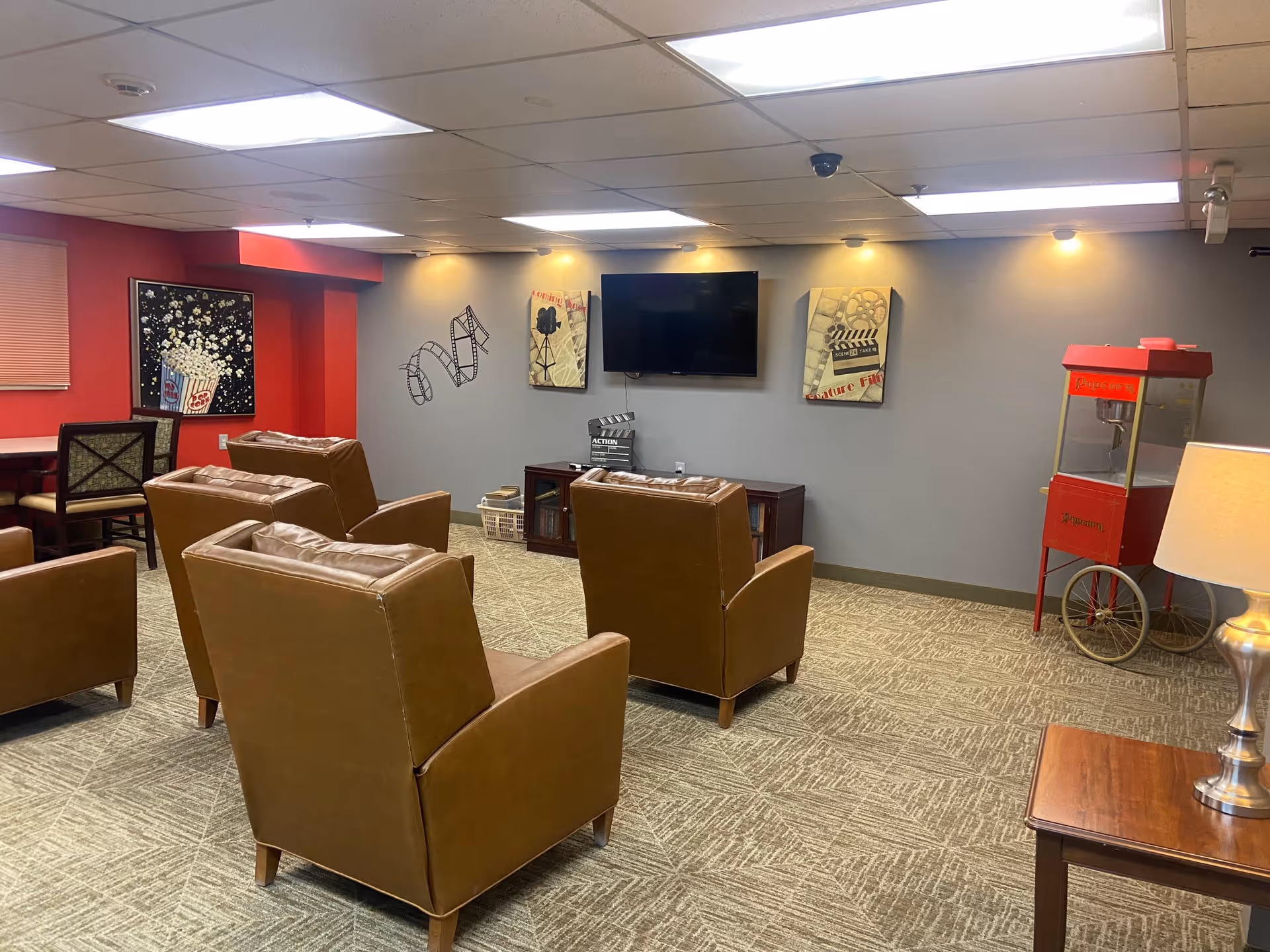 Seating area with brown armchairs facing a wall-mounted TV, wall art, and a red popcorn cart in a common room.