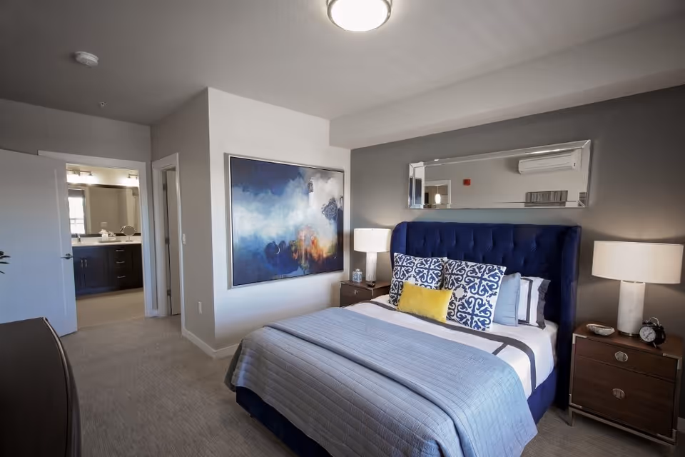 A modern bedroom with a blue upholstered bed featuring decorative pillows in blue and yellow. There are two wooden nightstands with lamps on either side of the bed. A large abstract painting hangs on the wall to the left, and a rectangular mirror is mounted above the bed. The room has a carpeted floor and neutral-colored walls. An open door reveals a bathroom with a vanity and mirror.